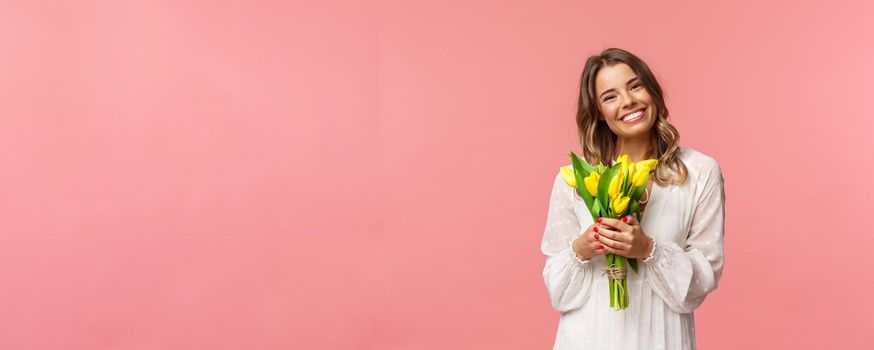 Holidays, beauty and spring concept. Portrait of lovely caucasian blond girl in white dress, smiling upbeat, holding yellow tulips, having perfect romantic date with flowers as gift, pink background.