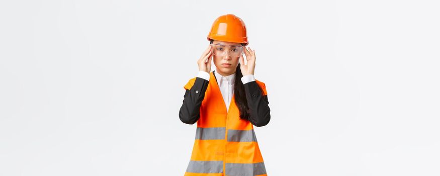 Serious-looking young asian female construction manager follow safety protocol, wearing protective glasses and helmet before entering enterprise, standing white background.