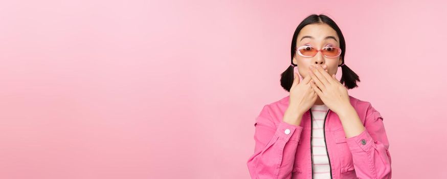 Portrait of shocked, stylish asian girl in sunglasses, closes mouth, looks with surprised face expression, stands over pink background. Copy space