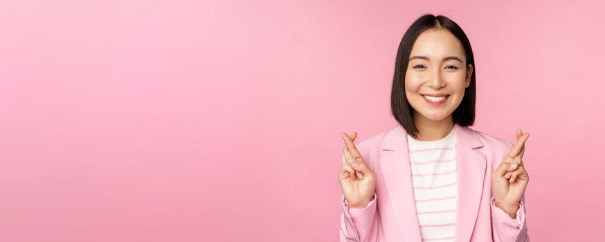 Happy asian businesswoman cross fingers for good luck, wishing, praying and hoping, smiling at camera, standing in suit over pink background.