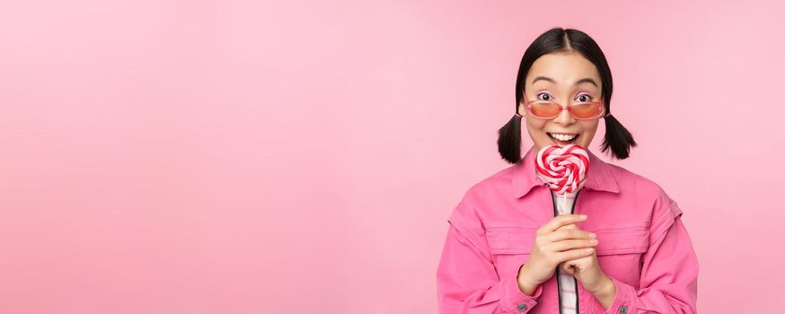 Silly and cute asian female model licking lolipop, eating candy sweet and smiling, looking excited, standing over pink background.