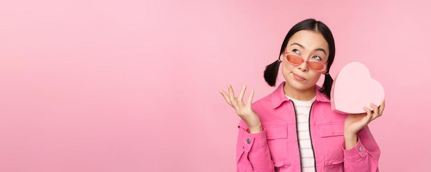 Image of stylish asian girlfriend in sunglasses, guessing whats inside gift box, heart-shaped present, standing over pink background.