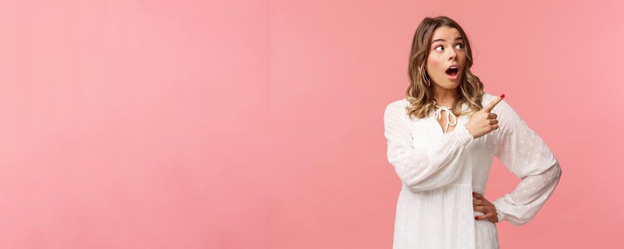 Surprised and excited good-looking blond girl in white cute dress, open mouth in amazement, look curious pointing upper right corner astonished, see something awesome, pink background.