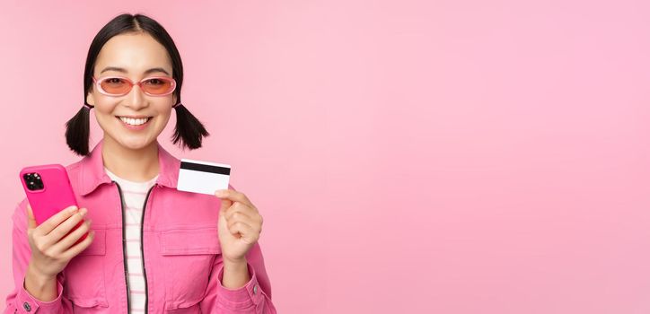 Online shopping. Smiling asian girl shopper, holding smartphone and credit card, paying in mobile app, standing over pink background.