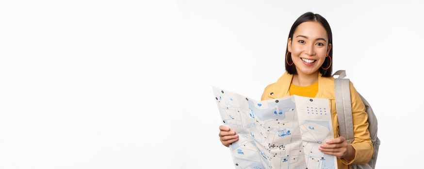 Tourism and backpacking. Smiling asian girl holding map and backpack, tourist searching way, travelling, standing over white background.