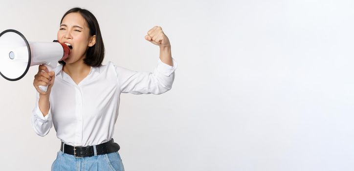 Enthusiastic asian woman, girl activist shouting at protest, using megaphone, looking confident, talking in loudspeaker, protesting, standing over white background.