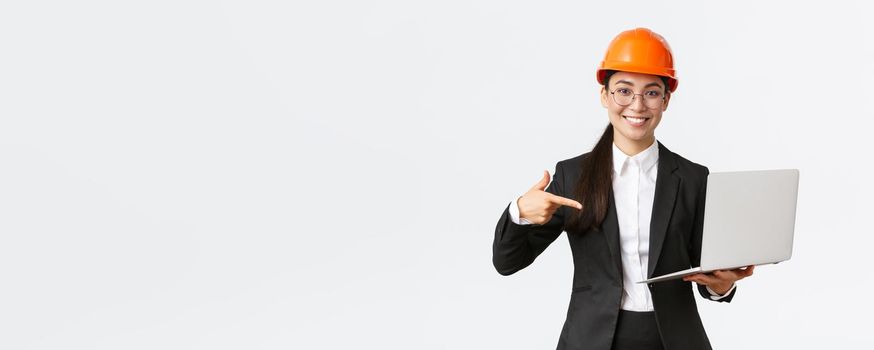 Smiling pleased asian female chief engineer showing chart with enterprise profit, making presentation during investors meeting, wearing safety helmet, pointing at laptop screen, white background.