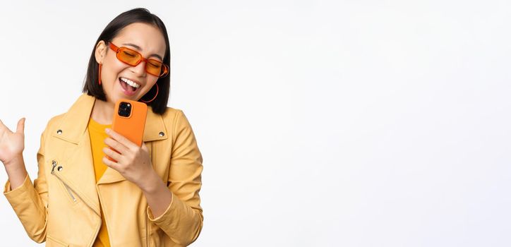 Technology and people concept. Happy dancing asian girl in sunglasses, using smartphone, singing and smiling, standing over white background.