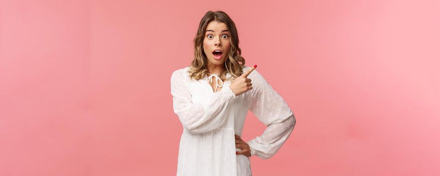 Surprised beautiful blond girl pointing upper right corner, gasping amused and staring at camera with fascinated expression, open mouth in awe to ask question about promo, pink background.