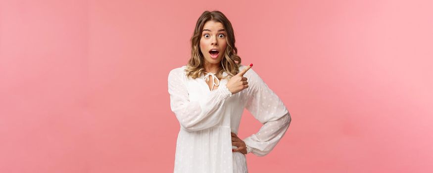 Surprised beautiful blond girl pointing upper right corner, gasping amused and staring at camera with fascinated expression, open mouth in awe to ask question about promo, pink background.