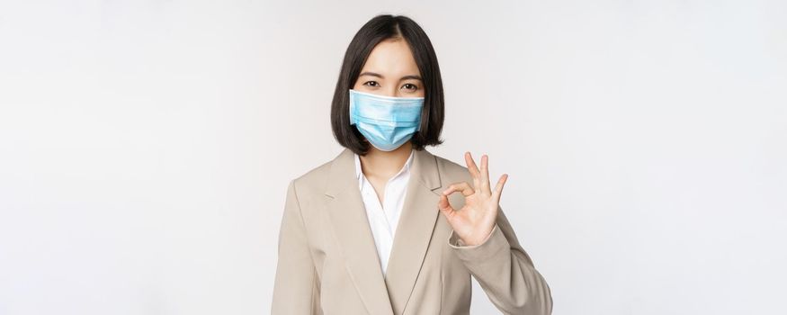 Coronavirus and workplace concept. Image of asian saleswoman, company worker in medical mask showing okay sign, smiling pleased, white background.