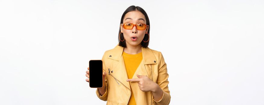 Portrait of stylish korean girl in sunglasses, smiling, pointing finger at smartphone screen, showing mobile phone application, standing over white background.