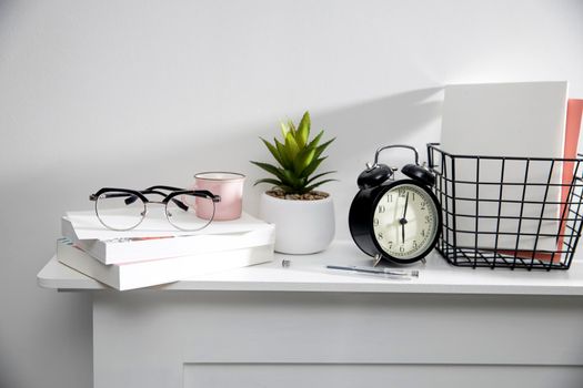 A stack of books, an artificial succulent plant in a ceramic pot, glasses, a metal basket with notebooks, a pen and a cup on a white table surface. Concept - office at home. Online training.