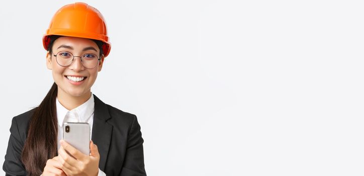 Close-up of successful female chief engineer, construction architect in safety helmet and business suit, glasses, looking at camera satisfied, smiling pleased as using mobile phone.