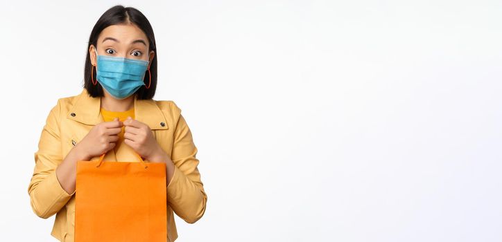 Covid-19 and shop concept. Young asian stylish woman, wearing medical face mask, holding shopping bag, going in malls during pandemic, white background.