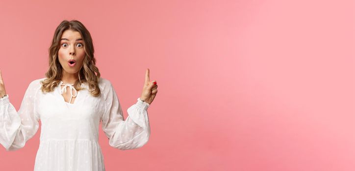 Waist-up portrait of amazed blond girl say wow in excitement, pointing fingers up, showing person top advertisement with cool special spring discount, standing pink background.