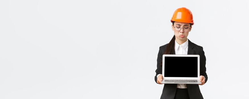 Disappointed gloomy asian female engineer showing bad results, wearing safety helmet and business suit, holding laptop computer and looking at screen displeased, standing white background pouting.