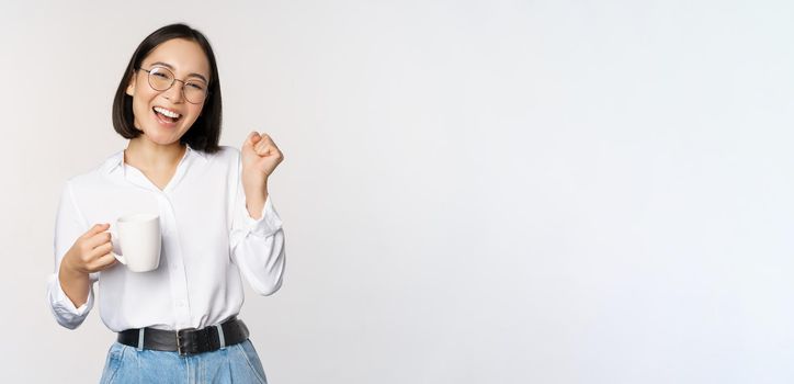 Happy dancing woman drinking coffee or tea from mug. Korean girl with cup, standing over white background.