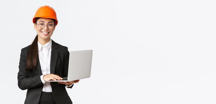 Smiling professional female saleswoman showing construction works on new house. Asian industrial engineer in safety helmet and business suit using laptop computer, enter data from factory.