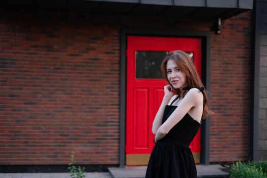 charming young woman in a black dress in front of modern building with red door. housewarming. buying a house. happy life in private house.