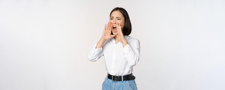 Image of young asian woman calling for someone, shouting loud and searching around, standing against white background.