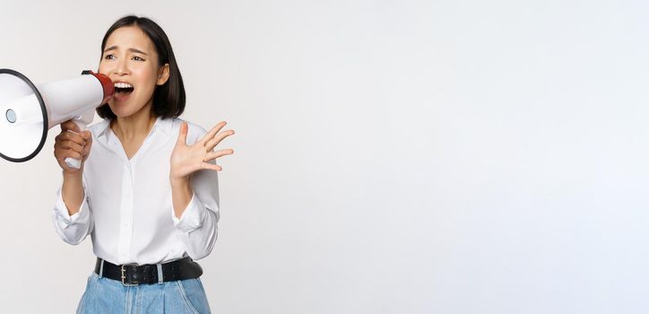 Image of young woman, korean activist, recruiter screaming in megaphone, searching, shouting at loudspeaker, standing over white background.