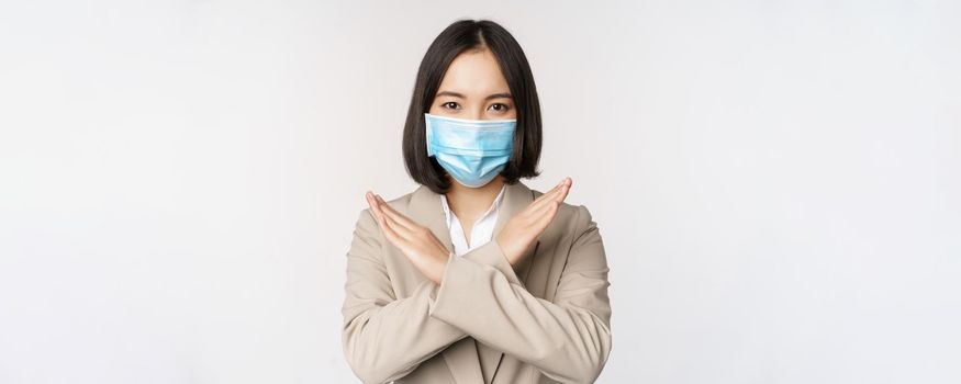 Coronavirus and workplace concept. Image of asian businesswoman, female in medical face mask shows stop, cross prohibit gesture, stands over white background.