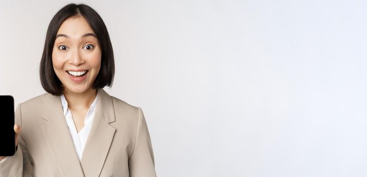 Image of asian corporate woman showing app interface, mobile phone screen, making surprised face expression, wow, standing over white background.