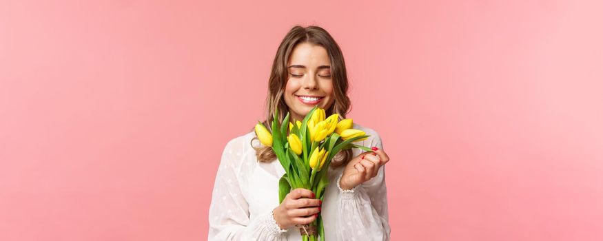 Spring, happiness and celebration concept. Close-up portrait of lovely, romantic blond girl sniffing smell of beautiful yellow tulips, close eyes and smiling happy, standing pink background.