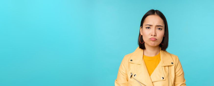 Close up of disappointed asian girl sulking, grimacing and frowning displeased, standing moody against blue background. Copy space