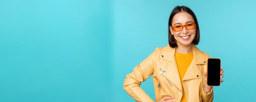 Image of young stylish asian female model in trendy sunglasses, showing mobile phone screen, application on smartphone interface, standing over blue background.