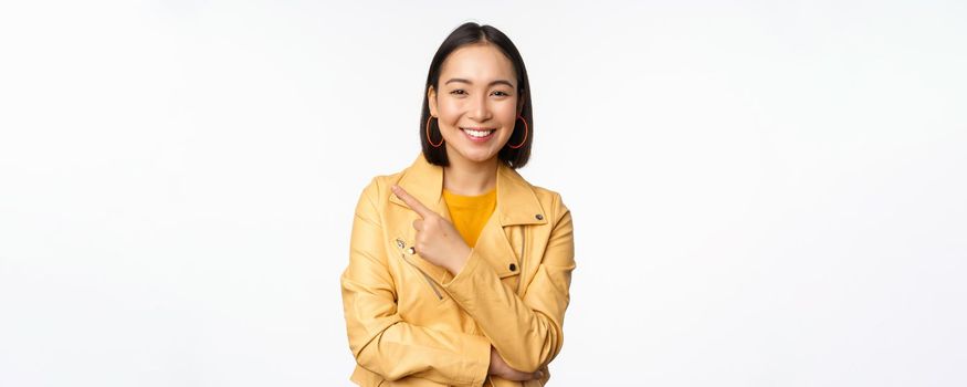 Portrait of smiling asian girl pointing finger left, showing logo or copy space, advertisement on empty side, standing over white background.
