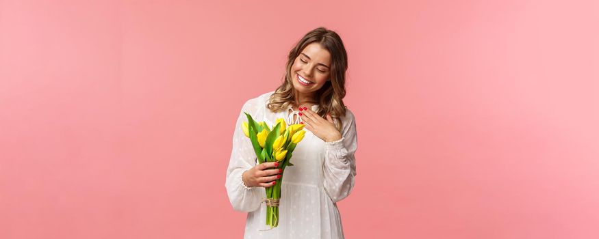 Holidays, beauty and spring concept. Portrait of pleased, cute romantic blond girl receive beautiful bouquet of flowers, looking at yellow tulips and touch heart grateful, smiling.