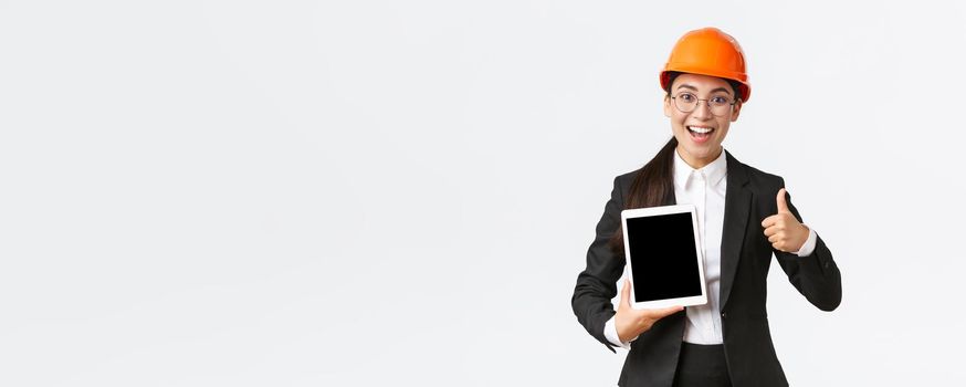 Impressed young female realtor, estate agent showing building during construction, wear safety helmet and business suit, holding digital tablet and make thumbs-up, recommend purchase.