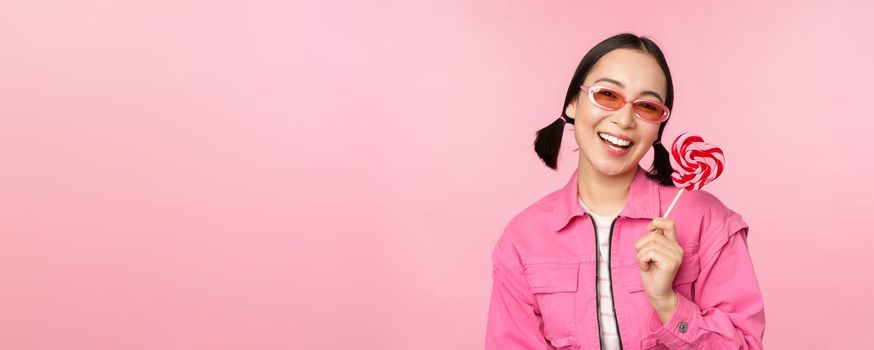 Stylish korean girl licking lolipop, eating candy and smiling, standing in sunglasses against pink background.