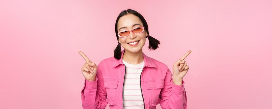 Choice. Stylish korean girl, asian female model points fingers sideways, shows two variants, product advertisement, demonstrating items, standing over pink background.