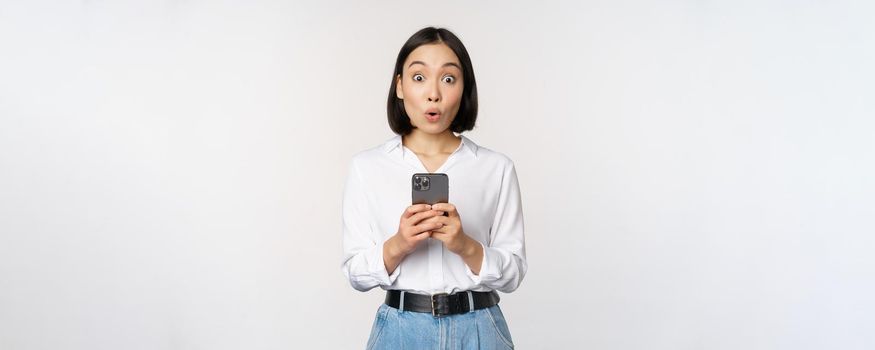 Technology concept. Portrait of asian woman with mobile phone, girl holding smartphone and reacting surprised at cellphone notification, app info, white background.