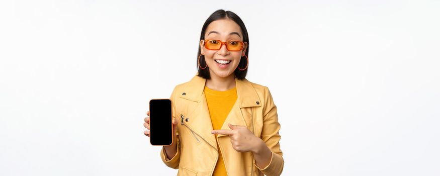 Portrait of stylish korean girl in sunglasses, smiling, pointing finger at smartphone screen, showing mobile phone application, standing over white background.