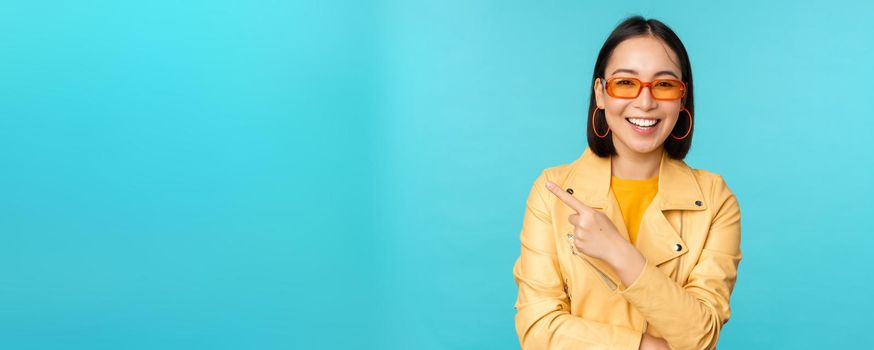 Smiling asian woman in sunglasses, pointing finger left, showing banner, logo or advertisement, standing over blue background. Copy space
