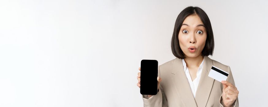 Corporate woman with happy, enthusiastic face, showing credit card and smartphone app screen, standing in suit over white background.