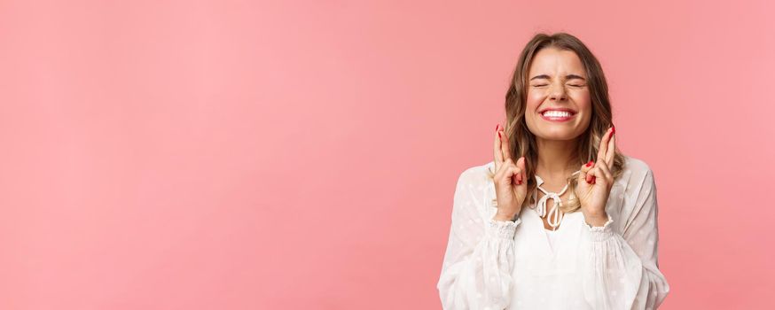 Portrait of excited hopeful blond girl making wish crossed fingers for good luck, close eyes and smiling putting all effort into pray, pleading for dream come true, anticipating over pink background.