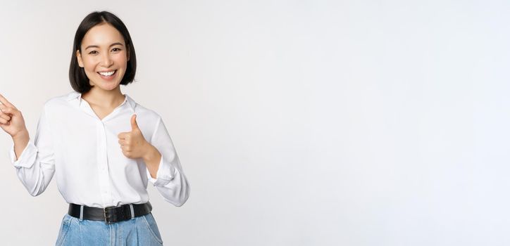 Image of young asian business woman, smiling while pointing finger left and showing thumbs up, recommending product, praise, standing over white background.