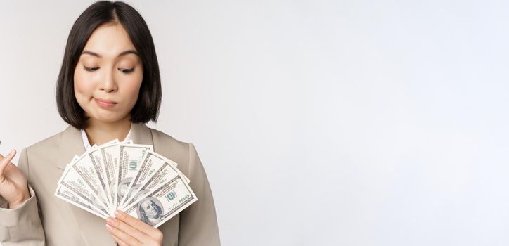 Image of asian corporate woman, happy businesswoman showing money, cash dollars and thinking, standing in suit over white background.