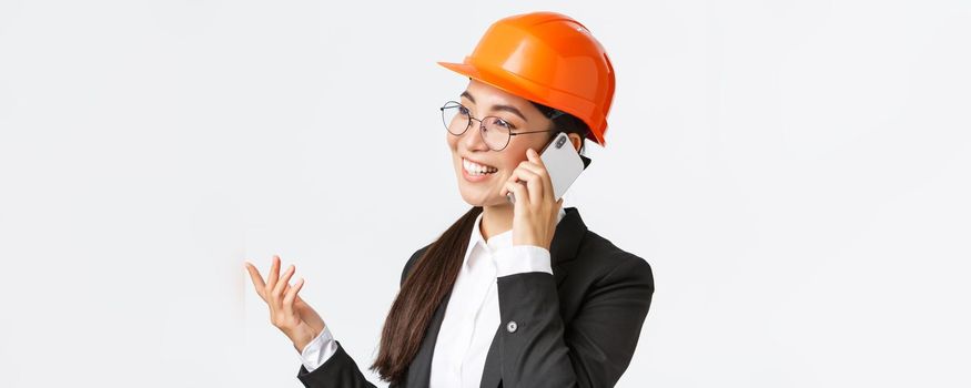 Close-up of professional smiling asian female entrepreneur at factory, chief engineer in safety helmet and suit, talking on phone, having business conversation with investors of enterprise.