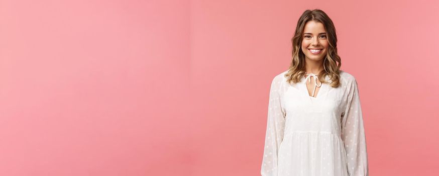 Spring, beauty and women concept. Portrait of tender feminine blonde girl with white beaming smile standing in light cute dress over pink background with happy attitude, positive emotions.