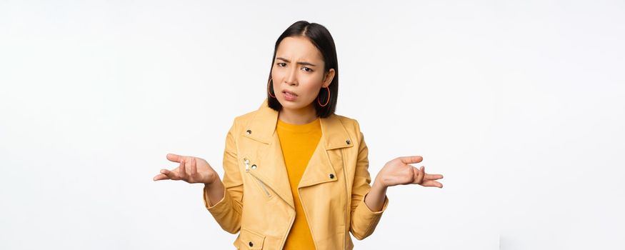 Image of confused young asian woman shrugging shoulders, looking puzzled and clueless at camera, cant understand, standing against white background.