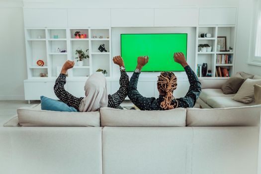 African Couple Sitting On Sofa Watching TV Together Chroma Green Screen Woman Wearing Islamic Hijab Clothes. High quality photo