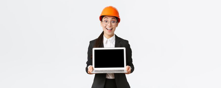 Excited happy asian female engineer, industrial manager in business suit and safety helmet showing laptop screen to investors or clients, amazed with great profit chart, introduce diagram.