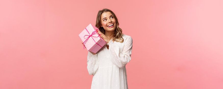 Holidays, celebration and women concept. Portrait of curious attractive young blond girl celebrating birthday, shaking pink gift box to found out whats inside, smiling intrigued look camera.