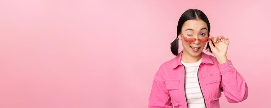 Eyewear advertisement. Stylish modern asian girl touches sunglasses, wears pink, poses against studio background. Copy space.